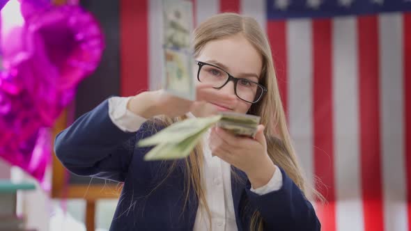 Rich Cute Schoolgirl Scattering Cash Looking at Camera Sitting in Classroom with American Flag at