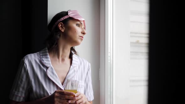 Woman in Striped Pajamas and with a Sleep Mask is Sitting on the Bed in the Bedroom and Drinking