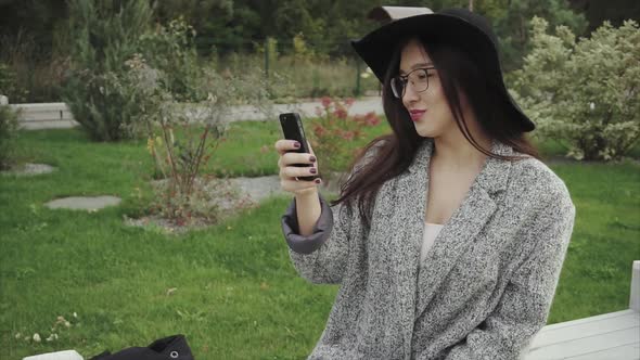 Young Attractive Woman Sits on Bench in the Park and Works on the Smartphone