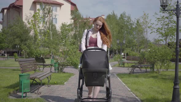 Young Happy Redhead Mother Walking with Baby Carriage and Smiling Along the Street on a Nice Spring