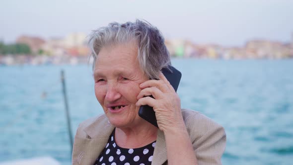 Old Woman Traveler Talks on Phone Standing on Lagoon Pier