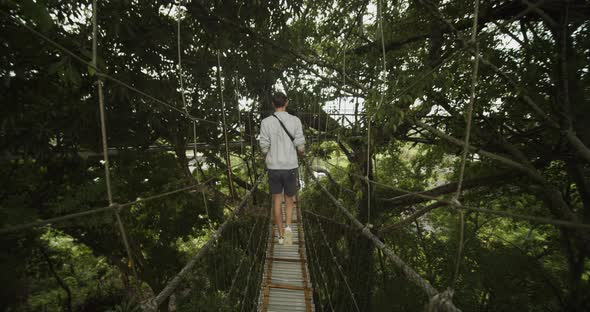 Dolly Shot Following a Young Man Carefully Walking Across a Rope Hanging Bridge Above a Tropical