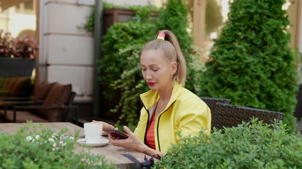 a Blonde in Red and Yellow Clothes is Sitting with a Phone and a Cup of Coffee at a Cafe Table