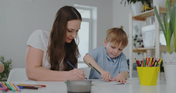 A Family of Two Children and a Young Mother Sitting at the Table Draws on Paper with Colored Pencils