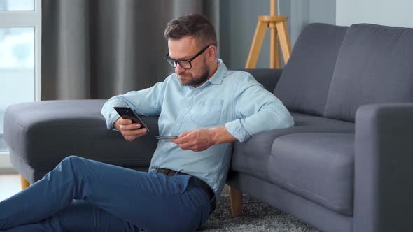 Bearded Man in Glasses Sits on the Carpet Near the Sofa and Makes Payment Online with a Credit Card