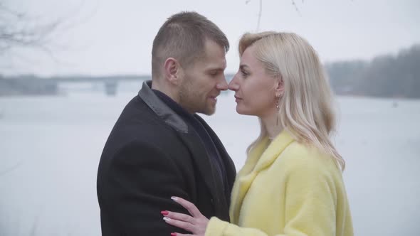 Happy Adult Couple Kissing, Hugging and Rubbing Noses As Standing on Riverbank on Cloudy Day