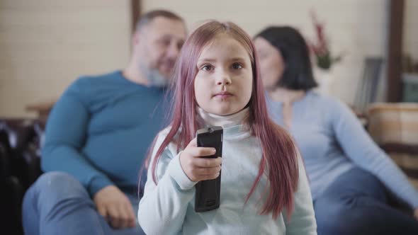 Portrait of Cute Girl Switching Channels with Remote Control As Blurred Father and Mother Sitting on