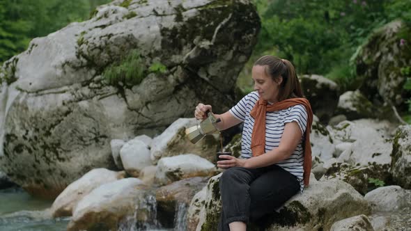 Smiling Woman pours coffee  from Mocha Pot against mountain river, road trip and staycation concept