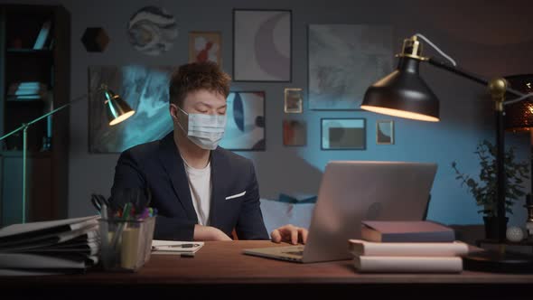 Young Asian Man Wearing Medical Mask Working Remotely at Home on Laptop Computer During Coronavirus