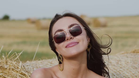 Brunette in Sunglasses with Blowing Hair Smiling at Camera in Sunny Hay Field