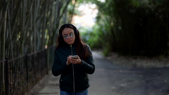 Adult Woman is Chatting Online in Smartphone and Listening to Music By Headphones Walk in Park