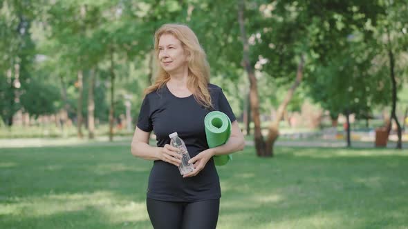 Portrait of Smiling Blond Mid-adult Woman Walking with Exercise Mat and Bottle of Water in Sunny