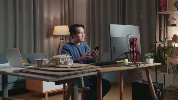 Asian Male Engineer With The House Model Using Smartphone While Working On A Desktop At Home