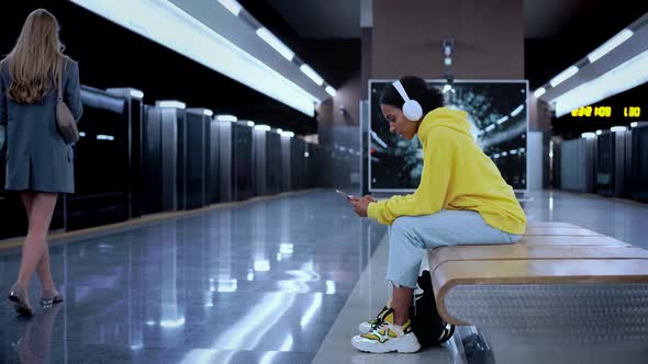 Young Girl Sitting in Modern Metro Station Waiting for Train Wearing Headphones