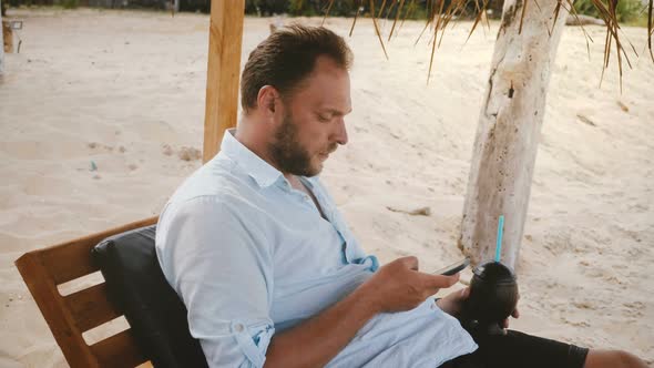 Happy Confident Male Tourist with Smart Watch and Fruit Cocktail Sitting in Beach Chair Using