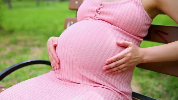 Happy Pregnant Woman Sitting on a Park Bench and Gently Stroking His Stomach with His Hand on a