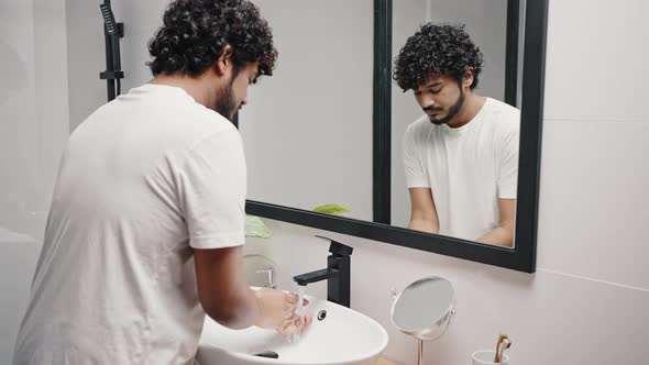 Young Indian Man Washes Hands Under Faucet in Bathroom