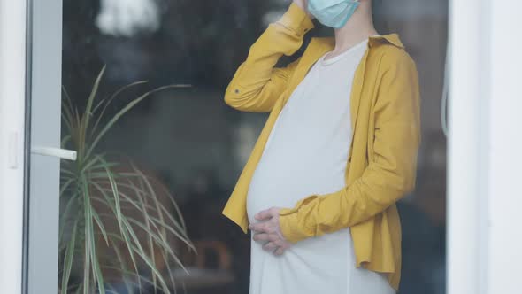 Unrecognizable Pregnant Woman in Covid Face Mask with Head Ache Standing Behind Glass Door Indoors