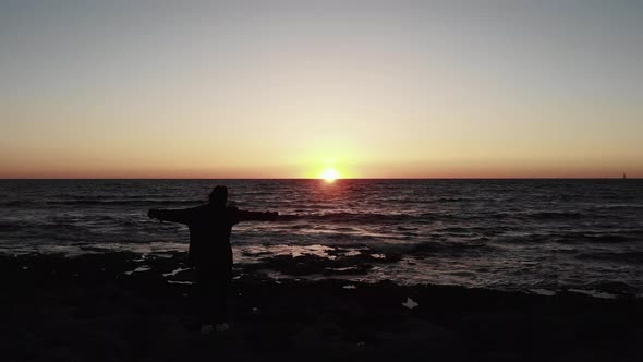 Silhouette of Young Woman Standing on The Rocky Beach with Hands Spread to Sides on Sunset at Sea