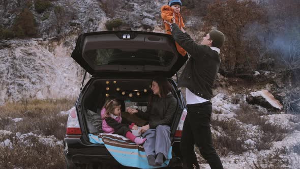 Traveler Family  sitting in the open trunk of a black car with kids near bonfire