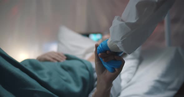 Doctor in Gloves and Ppe Suit Holding Hands of Sick Covid19 Aged Lady Lying in Bed