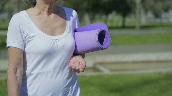 Middle-aged Female Body Walking in Park with Yoga Mat in Hand