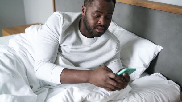 Sleepy African man texting by phone in bed