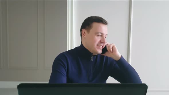 Happy young businessman in blue sweater talking on mobile phone while working on laptop 