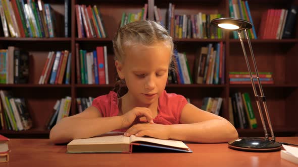 Six Year Old Girl Reading Sitting at the Table By the Light of a Desk Lamp