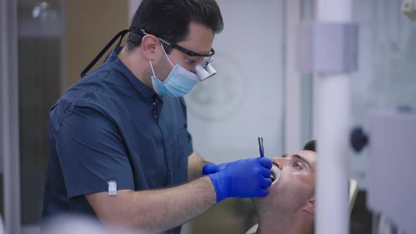 Side View Focused Middle Eastern Man in Dental Binocular Loupes Examining Teeth of Patient in