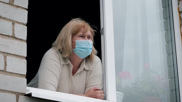 Mature Woman In A Protective Medical Mask Looks Out Of The Window At The Street