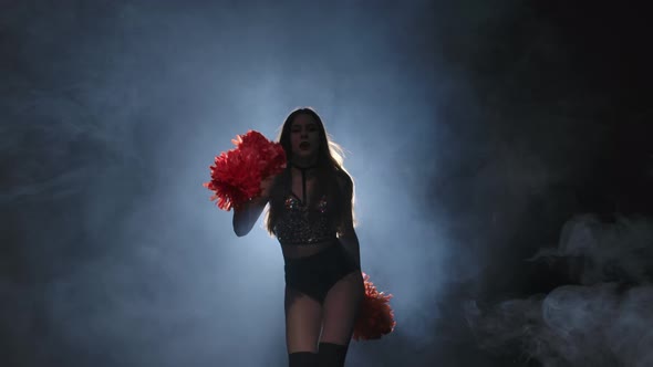 Young Female Cheerleader with Orange Pompoms in Uniform is Dancing on Black Smoky Studio Background