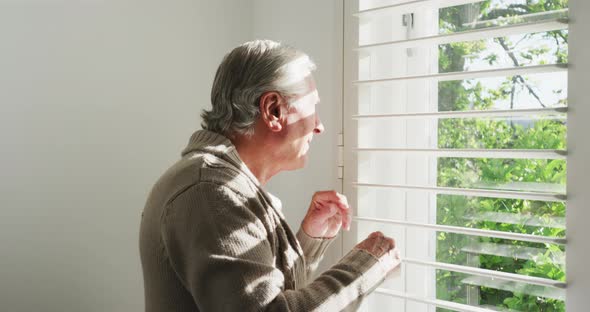 Retired senior man in cardigan sweater looking out through window blinds at home