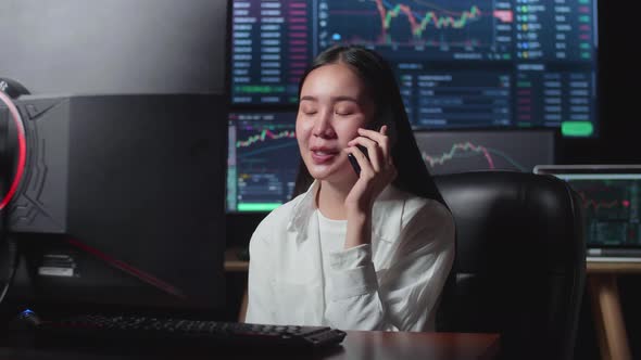 Asian Young Female Stock Market Broker Talking On Mobile Phone With Analyzing Graphs At Background