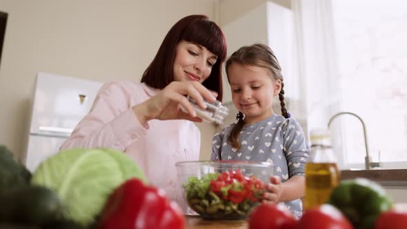 Portrait of Smiling Young Mom i and Little Daughter Adding Salt to the Salad While Cooking in