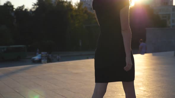 Young Unrecognizable Girl in Black Dress Walking Along Urban Street at Sunset Time