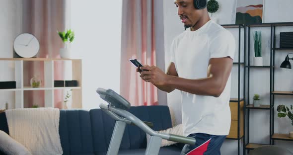 Black-Skinned Guy in Headphones Walking on Running Track at Home and Applying his Phone