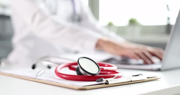 Red Stethoscope on Table in Background Doctor is Typing on Laptop