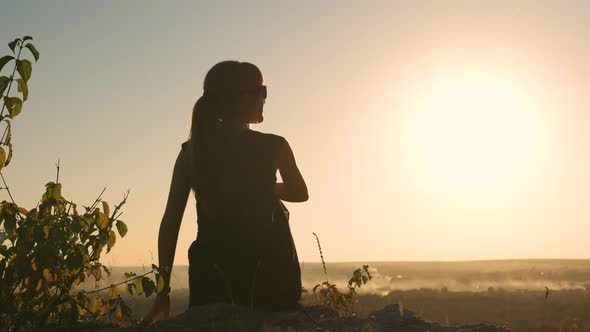 Young slim woman in black short dress sitting on a rock relaxing outdoors at summer sunset.