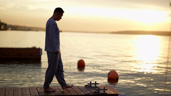 Young Sailor is Enjoying Beautiful Surise Over Sea Walking on Wooden Pier and Sitting on Edge