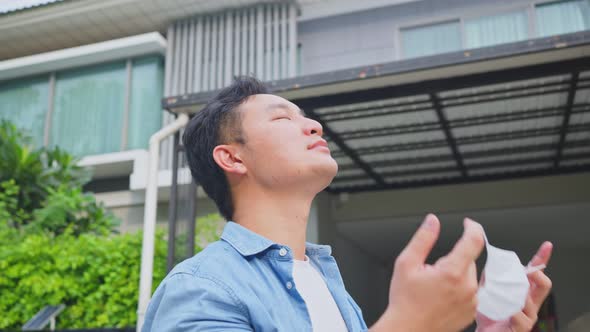 Asian attractive man stand in front of his house removing protective face mask and take deep breath.