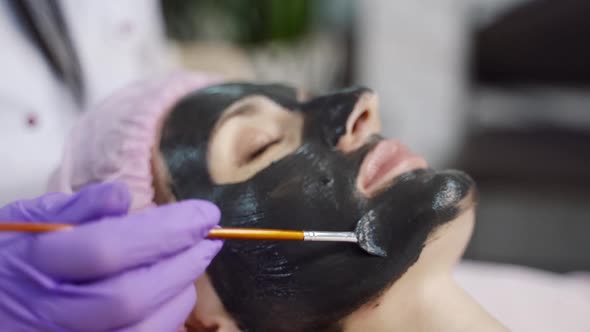 A specialist is spreading mask with brush on the face of the woman in the spa salon.