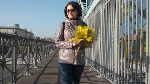 A Middleaged Brunette Woman in Black Glasses Walks Alone with a Bouquet of Yellow Leaves on the