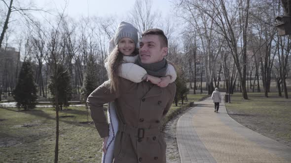 Portrait of Happy Caucasian Young Father Holding Little Pretty Daughter on Back. Cheerful Girl and