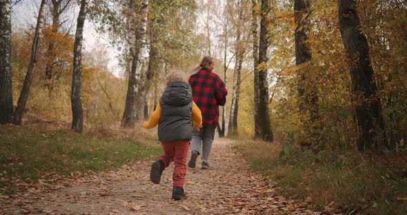 Young Woman and Little Child Are Running in Autumn Forest Over Walkpath, Son Is Following Mother