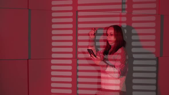 Young Woman with Mobile Phone Standing Near Wall Highlighting with Shadows of Jalousie