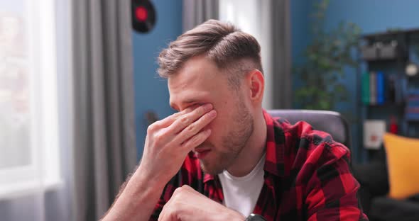 A Handsome Young Man Rubs His Eyes with Fatigue While Working Remotely From Home