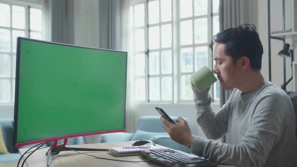 Asian Man With Green Screen Computer Using Mobile Phone And Drinking Coffee While Working At Home