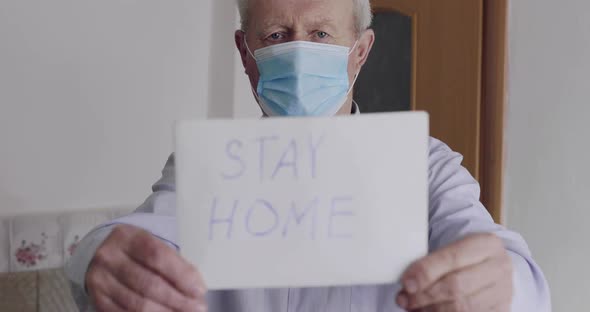 Worrid Senior Man in Mask on Isolation Shows at Camera Inscription "STAY HOME"