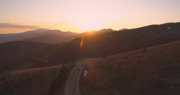 Drone Following Lonely Car Driving on Curved Mountain Road Reflecting the Sunset Lights